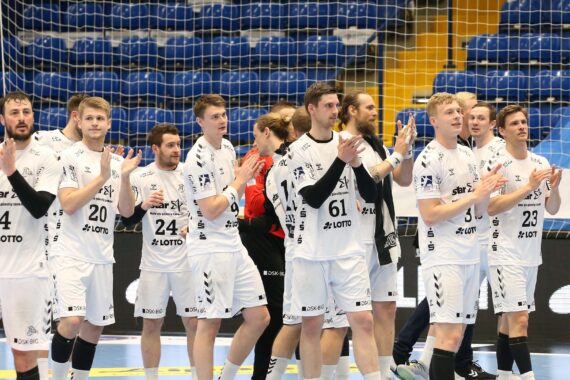 Eine Männerhandballmannschaft in weißen Trikots steht gemeinsam auf einem Hallenplatz, klatscht und blickt zum Publikum. Die Spieler scheinen das Publikum zu grüßen, während im Hintergrund ein Netz und leere blaue Sitze zu sehen sind.
