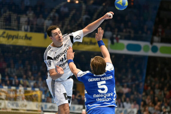Ein Handballspieler in einem weißen Trikot springt und wirft den Ball, während ein Verteidiger in einem blauen Trikot versucht, ihn während eines Hallenspiels zu blocken, wobei im Hintergrund Zuschauer zu sehen sind.