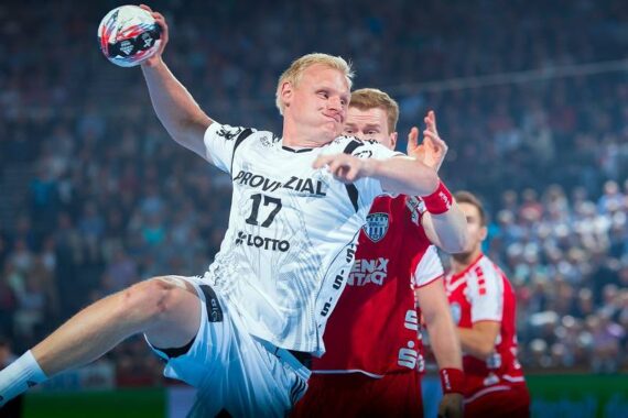 Ein männlicher Handballspieler in einem weißen Trikot springt, um den Ball zu werfen, während ein Verteidiger in einem roten Trikot versucht, ihn während eines intensiven Spiels in einer überfüllten Halle zu blockieren.