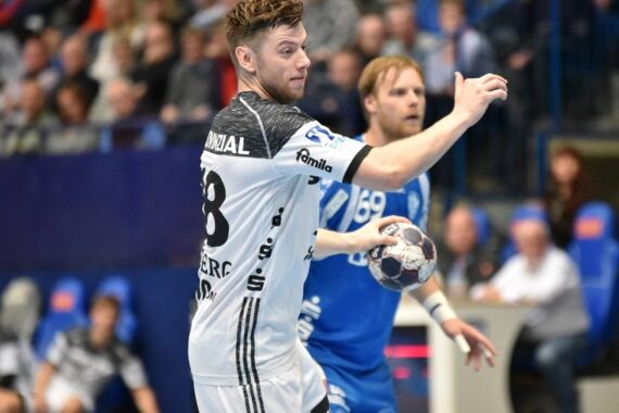 Ein männlicher Handballspieler in einem weiß-schwarzen Trikot hält den Ball und gestikuliert mit der Hand, während ein Gegner in einem blau-weißen Trikot während eines Spiels in einer Halle hinter ihm steht.