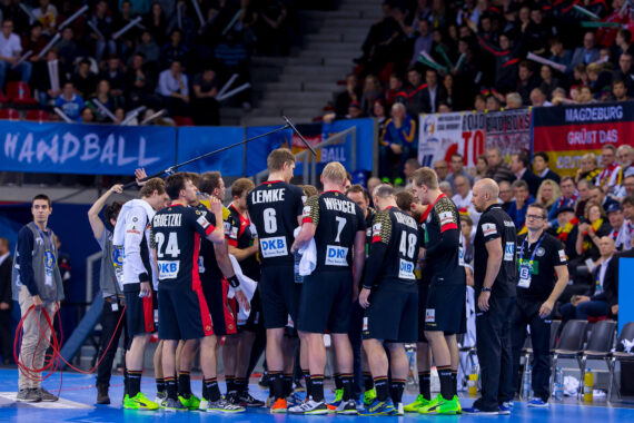 Eine Männerhandballmannschaft in schwarzen Trikots versammelt sich während eines Spiels in einem Huddle, während Trainer und Betreuer in der Nähe stehen. Im Hintergrund sind Zuschauer mit Bannern und Fahnen zu sehen.
