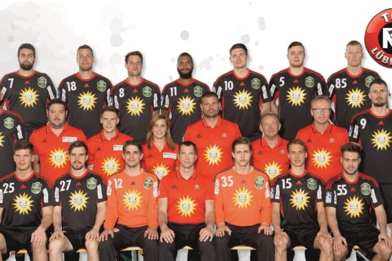 Gruppenfoto der Handballmannschaft des TuS N-Lübbecke in schwarz-orangenen Trikots mit gelben Sonnenlogos, die in drei Reihen mit dem Teamstab posieren. Das Wappen des TuS N-Lübbecke befindet sich in der oberen rechten Ecke.