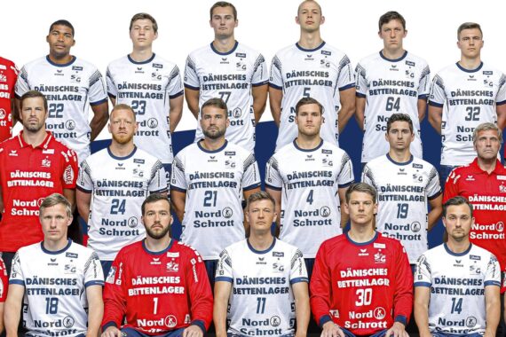 Eine Männer-Handballmannschaft stellt sich für ein Gruppenfoto auf. Die meisten Spieler tragen weiße und blaue Trikots, andere tragen rote. Sie stehen und sitzen in Reihen und blicken direkt in die Kamera, vor einem schlichten Hintergrund.