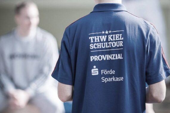 Eine Person, die ein blaues Hemd mit der weißen Aufschrift THW Kiel Schultour Provinzial Förde Sparkasse auf dem Rücken trägt, steht im Mittelpunkt, während eine andere Person in heller Kleidung unscharf im Hintergrund sitzt.