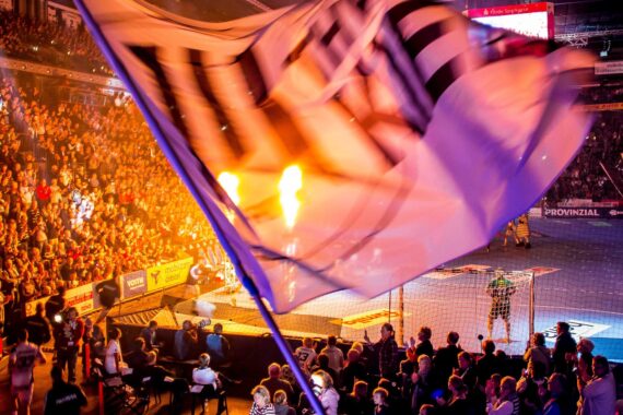 Eine große Flagge weht im Vordergrund, während eine vollbesetzte Halle ein Handballspiel verfolgt. Helle Flammen und eine dramatische Beleuchtung sorgen für eine lebhafte Atmosphäre.