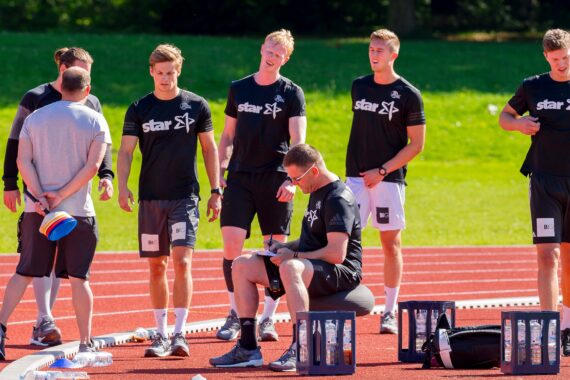 Eine Gruppe von sechs Männern in Sportkleidung versammelt sich auf einer Laufbahn, einige stehen, einer sitzt mit einem Klemmbrett. In der Nähe stehen Wasserflaschen und Kisten, und im Hintergrund ist hellgrünes Gras zu sehen.