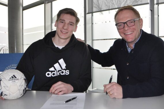 Zwei Männer sitzen in einem Haus an einem Tisch. Der jüngere Mann trägt einen Adidas-Kapuzenpulli und hält einen Fußball, während der ältere Mann mit Brille einen Stift in der Hand hält. Auf dem Tisch liegt ein Dokument, das auf eine Unterzeichnung oder Vereinbarung hindeutet. Beide lächeln.