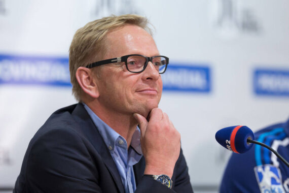 Ein Mann mit kurzen blonden Haaren und Brille, der einen Anzug trägt, sitzt an einem Tisch mit einem Mikrofon vor sich, lächelt und stützt sein Kinn auf seine Hand während einer Pressekonferenz.
