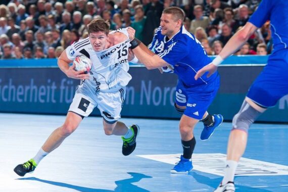 Ein männlicher Handballspieler in einem weißen Trikot versucht, sich mit dem Ball in der Hand zu befreien, während ein Verteidiger in einem blauen Trikot ihn während eines Spiels am Arm festhält, während im Hintergrund eine Menschenmenge zusieht.