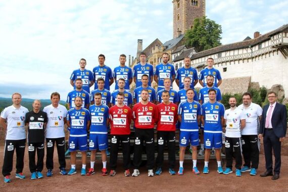 Eine Handballmannschaft der Männer posiert in Uniform im Freien, mit Trainern und Betreuern im Vordergrund. Dahinter befindet sich ein historisches Gebäude mit einem Turm, umgeben von Grünflächen unter einem teilweise bewölkten Himmel.