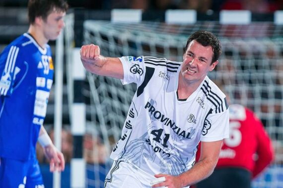 Ein Handballspieler in einem weißen Trikot mit der Nummer 41 jubelt auf dem Spielfeld und ballt die Faust, während ein anderer Spieler in einem blauen Trikot in der Nähe steht und im Hintergrund ein Torwart zu sehen ist.