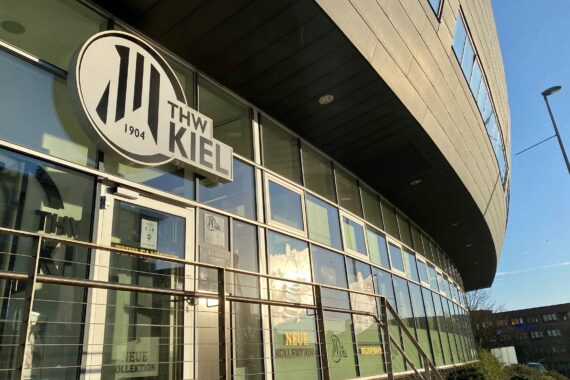 Das Äußere eines modernen Gebäudes mit großen Glasfenstern, auf denen sich das Logo des Handballvereins THW Kiel und der Schriftzug über dem Eingang spiegeln, reflektiert das Sonnenlicht und den klaren blauen Himmel.