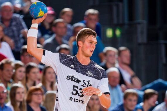 Ein männlicher Handballspieler in einem weiß-schwarzen Trikot bereitet sich während eines Spiels auf den Wurf eines blauen Handballs vor, während im Hintergrund eine Zuschauermenge zusieht.