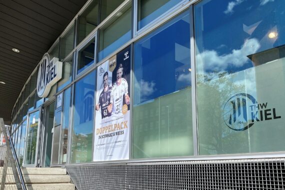 Die Außenansicht des THW Kiel mit großen Fenstern, einem Plakat mit zwei Sportlern und dem Logo des THW Kiel auf der Glasscheibe. Eine Treppe und ein Metallgeländer führen zum Eingang.
