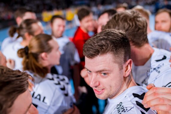 Eine Gruppe von Sportlern in weißen Trikots drängt sich zusammen, wobei ein Spieler im Mittelpunkt steht und leicht zur Seite schaut. Der Hintergrund ist unscharf, was die Geschlossenheit und Konzentration der Mannschaft unterstreicht.