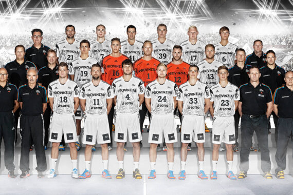 Eine Profi-Handballmannschaft steht in zwei Reihen für ein Gruppenfoto, die Spieler in weißen Trikots, die Betreuer in Schwarz. Der Hintergrund zeigt eine jubelnde Menge in einem Stadion unter hellen Lichtern.