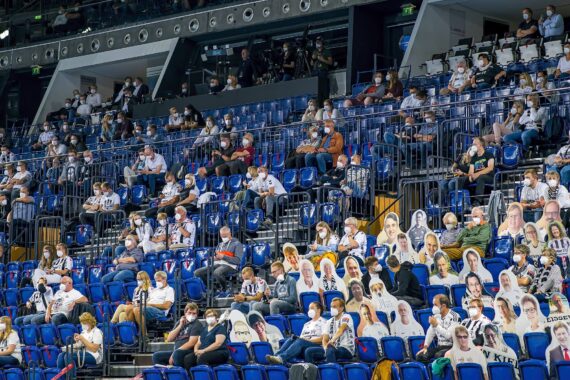 Die Fans sitzen in einem Stadion weit voneinander entfernt, viele tragen Masken. Einige Sitze sind mit Pappfiguren gefüllt, um den Anschein einer größeren Menschenmenge zu erwecken und die Maßnahmen zur sozialen Distanzierung während einer Sportveranstaltung zu unterstreichen.