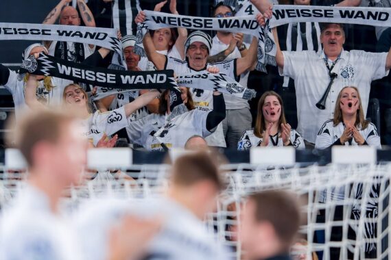 Eine Gruppe begeisterter Sportfans in schwarz-weißen Trikots hält WEISSE WAND-Schals hoch und jubelt bei einem Sportereignis leidenschaftlich hinter einem Netz.