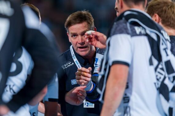 Ein Trainer spricht während einer Auszeit aufmerksam zu seiner Mannschaft, umgeben von Spielern in schwarzen und weißen Uniformen auf einem Sportplatz.