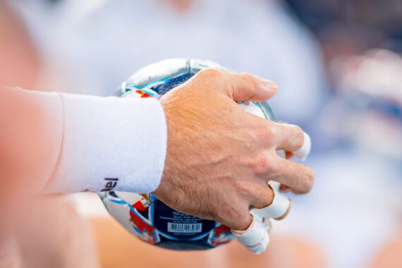 Nahaufnahme einer Hand, die ein weißes Sportarmband und Fingertape trägt und einen bunten Handball vor einem unscharfen Hintergrund hält.