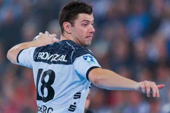Ein männlicher Handballspieler in einem weiß-schwarzen Trikot mit der Nummer 18 ist in Aktion und bereitet sich darauf vor, den Ball während eines Spiels zu werfen. Der Hintergrund zeigt eine unscharfe Menschenmenge in einer Sportarena.
