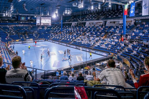 Eine große Halle mit blauen Sitzen und einem hellblauen Handballfeld, teilweise gefüllt mit sozial distanzierten Zuschauern, die ein Handballspiel live verfolgen. Von der Decke hängen Transparente und helle Lichter.