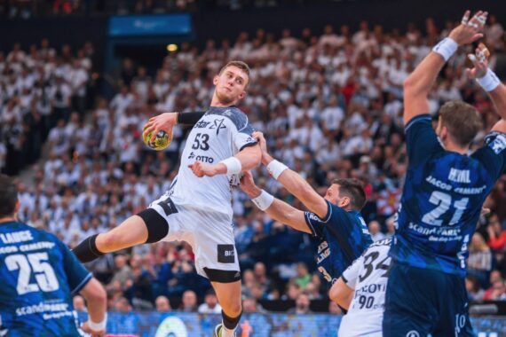 Ein Handballspieler in einem weißen Trikot springt in die Luft, um den Ball zu werfen, während zwei Gegner in blauen Trikots versuchen, ihn während eines intensiven Spiels in einer überfüllten Arena zu blockieren.