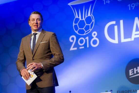 Ein Mann in einem braunen Anzug hält Notizen und spricht auf der Bühne einer Sportveranstaltung. Hinter ihm ist ein großer blauer Hintergrund mit einem Handball-Logo, "2018" und "Glasgow" sowie den Logos der EHF und der Europäischen Handballföderation zu sehen.