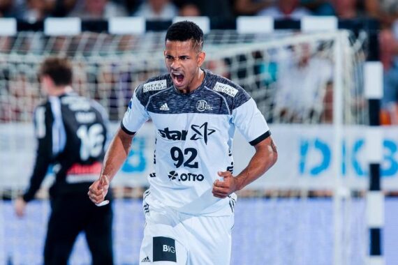 Ein männlicher Handballspieler in einem weiß-grauen Trikot mit der Nummer 92 jubelt energisch auf dem Spielfeld, ballt die Fäuste und schreit, während im Hintergrund ein Tornetz und ein anderer Spieler zu sehen sind.