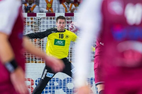 Ein Handballtorwart in einem gelben Trikot bereitet sich darauf vor, einen Schuss abzublocken, während die gegnerischen Spieler in kastanienbraunen Trikots im Vordergrund unscharf zu sehen sind.