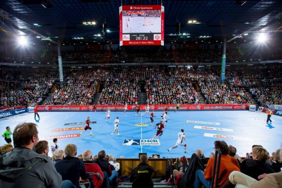 Eine überfüllte Halle mit Zuschauern, die ein Handballspiel auf einem blauen Spielfeld verfolgen; Spieler in weißen und roten Trikots treten gegeneinander an, und über dem Spielfeld hängt eine große Anzeigetafel, die den Spielstand anzeigt.