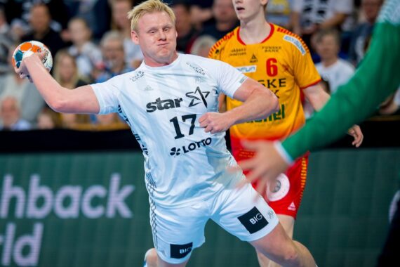 Ein männlicher Handballspieler in einem weißen Trikot bereitet sich auf den Wurf vor, während im Hintergrund ein Gegner in einem rot-gelben Trikot und ein Torwart in Grün zu sehen sind.
