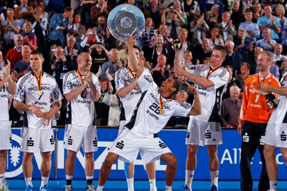 Eine jubelnde Handballmannschaft in weißen Trikots feiert auf einem Spielfeld, während ein Spieler eine große Trophäe in die Höhe reckt. Die Mannschaftskameraden jubeln und klatschen, während die Menge im Hintergrund applaudiert.