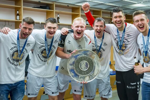 Sechs jubelnde männliche Athleten mit Medaillen und Mannschaftshemden feiern in einer Umkleidekabine, halten eine große silberne Trophäenplatte in der Hand und legen lächelnd die Arme umeinander. Im Hintergrund sind Flaschen zu sehen.