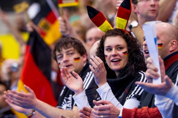 Eine Gruppe begeisterter Sportfans, viele mit schwarzen, roten und gelben Accessoires und Gesichtsbemalung, jubelt auf der Tribüne. Eine Frau in der Mitte trägt ein Stirnband mit den Farben der deutschen Flagge und lächelt.