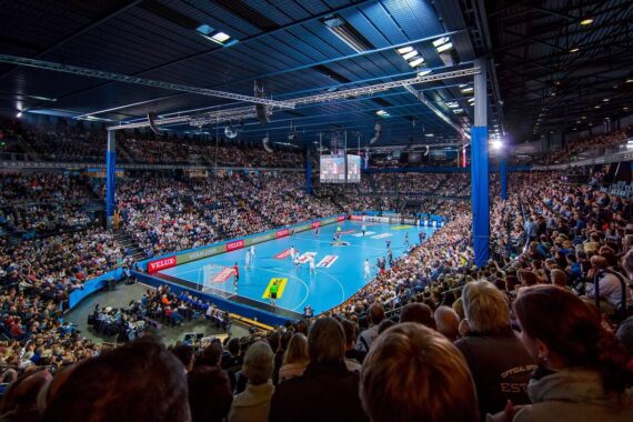 Eine große Halle, gefüllt mit Zuschauern, die ein professionelles Handballspiel auf einem blauen Spielfeld verfolgen, mit heller Beleuchtung und mehreren von der Decke hängenden Anzeigetafeln.