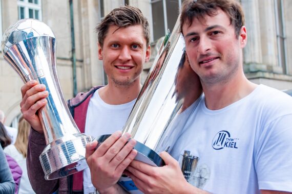 Zwei Männer in weißen Hemden stehen draußen und lächeln in die Kamera, während sie jeweils eine große silberne Trophäe halten. Im Hintergrund sind Menschen und ein Gebäude zu sehen.