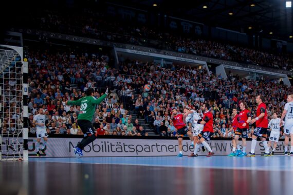 Ein Handballspiel in einer überfüllten Halle. Spieler in rot-weißen Trikots zielen auf das Tor, während ein Torwart in Grün sich darauf vorbereitet, den Ball zu blocken. Die Zuschauer füllen die Tribünen im Hintergrund.