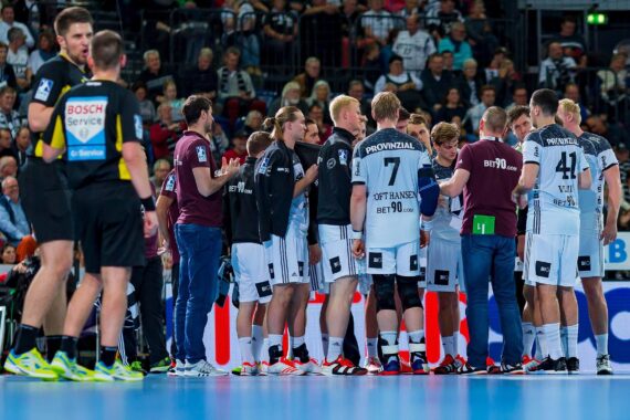Eine Handballmannschaft versammelt sich während einer Auszeit auf dem Spielfeld, umringt von Trainern und Betreuern, mit den Schiedsrichtern in der Nähe und den Zuschauern auf der Tribüne.