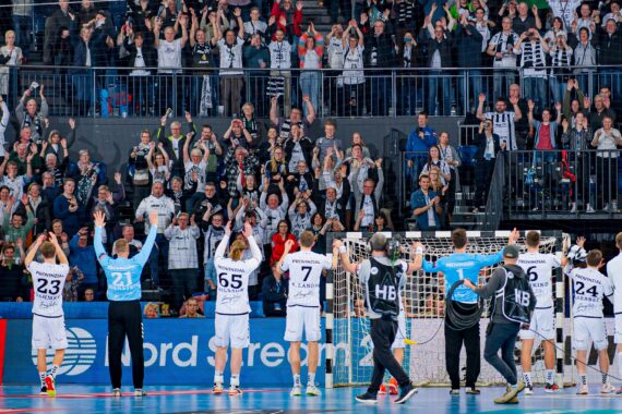 Handballspieler auf einem Spielfeld heben jubelnd die Arme vor einer jubelnden Menge, die ebenfalls hinter einer Absperrung in einem Stadion steht und klatscht.