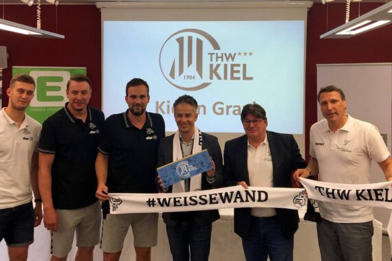 Sechs Männer stehen in einer Halle vor einer Projektion des THW Kiel-Logos, zwei halten einen THW Kiel #WEISSEWAND-Schal, ein anderer eine blaue Box. Einige tragen Markenshirts. Sie schauen lächelnd in die Kamera.