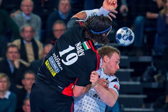 Zwei männliche Handballspieler in Aktion, einer in einem schwarzen Trikot blockt einen Spieler in einem weißen Trikot, während der Ball zwischen ihnen hindurchfliegt, während die Zuschauer im Hintergrund zusehen.