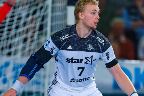 Ein männlicher Handballspieler, der ein weißes und schwarzes Trikot mit der Nummer 7 und Sponsorenlogos trägt, steht während eines Spiels in einer Action-Pose. Er trägt eine Armschiene am rechten Arm und ist auf das Spiel konzentriert.