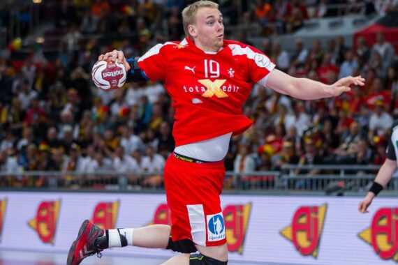 Ein Handballspieler in einem roten Trikot springt mit dem Ball in der rechten Hand in die Luft, um ihn zu werfen. Sein Hemd ist hochgezogen und zeigt seinen Oberkörper. Eine Menschenmenge schaut von der Tribüne im Hintergrund zu.