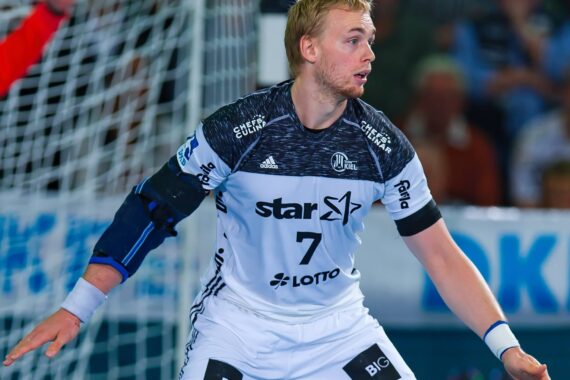 Ein männlicher Handballspieler, der ein weiß-schwarzes Trikot mit der Nummer 7 trägt, bereitet sich während eines Spiels mit ausgestreckten Armen und konzentriertem Blick auf die Verteidigung vor.