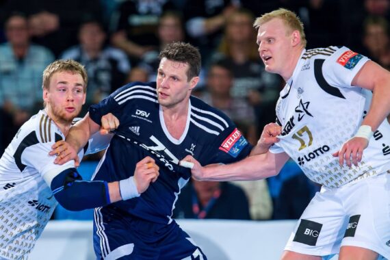 Drei männliche Handballspieler in Aktion während eines Spiels. Zwei Spieler in weißen Trikots versuchen, einen Spieler in einem marineblauen Trikot, der den Ball hält, zu blockieren, während im Hintergrund eine Menschenmenge zusieht.