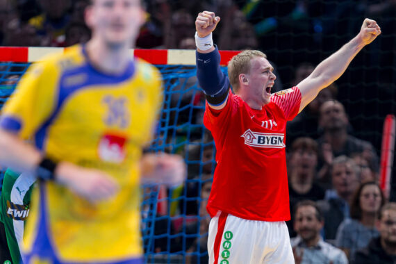 Ein Handballspieler in einem roten Trikot jubelt mit erhobenen Fäusten, während ein unscharfer Spieler in einem gelben Trikot im Vordergrund steht. Im Hintergrund schauen Zuschauer aufmerksam zu.