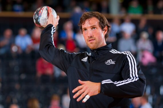 Ein männlicher Handballspieler in einem schwarzen Trainingsanzug bereitet sich während eines Spiels oder Trainings auf den Wurf vor, wobei die Zuschauer im Hintergrund unscharf zu erkennen sind.