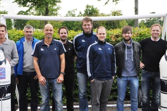 Acht Männer stehen im Freien vor einem Fußballtor und Sträuchern und posieren für ein Gruppenfoto. Zwei aufblasbare Handball-Trainingsattrappen mit Cartoon-Gesichtern sind an beiden Enden der Gruppe positioniert.