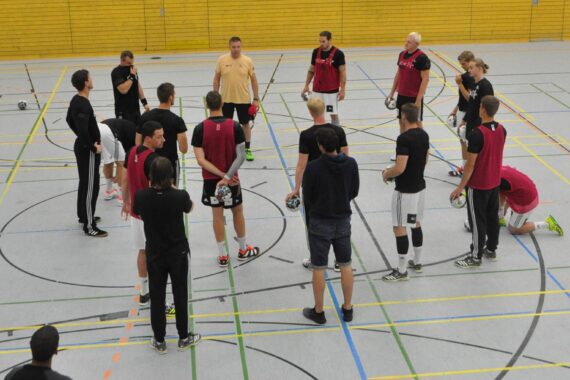 Eine Gruppe von Menschen in Sportkleidung steht in einem Kreis auf einem Hallenplatz und hört einem Trainer in einem gelben Hemd zu. Einige halten Handbälle, und der Boden des Platzes ist mit bunten Linien markiert.